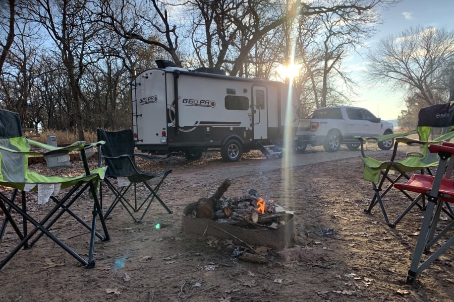 Sunny RV rental camp setup with a Geo Pro travel trailer, pickup truck, chairs, and a warm campfire in the woods.