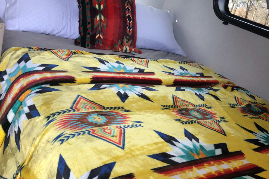Inside an RV rental: a bed with bright yellow blanket & patterned pillows, offering serene nature views from its window.