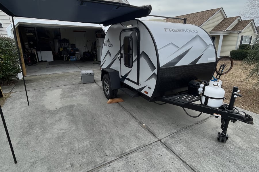 A FREESOLO teardrop RV rental, with an extended awning and propane tank, rests on a driveway beside a house.