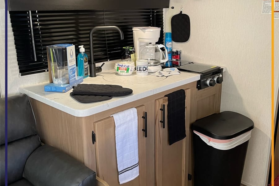 Compact rv rental kitchen with sink, coffee maker, microwave, stove, and cabinets, plus a cozy sofa and a colorful rug.