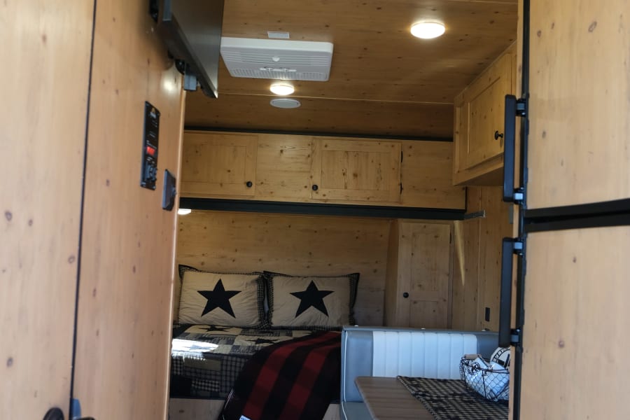 Cozy RV rental interior: wood-paneled walls, bed with star pillows, dinette, TV, and fridge.