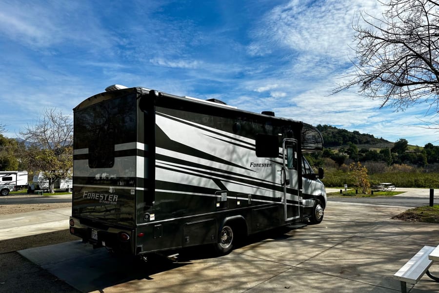 A large black and white Forester RV rental is parked on pavement at a sunny campground, surrounded by trees and hills.
