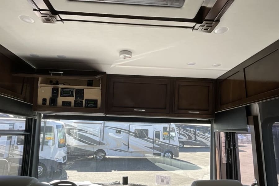 Inside an RV rental: overhead control panel and dark wood cabinets are visible from the driver's seat, with other RVs outside.