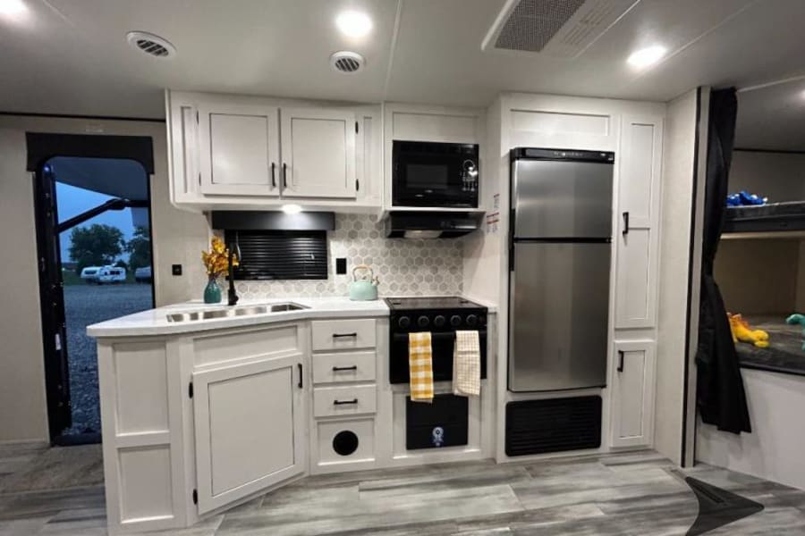 Modern RV rental kitchen: light cabinets, stainless steel appliances, sink, stove, and bunk beds. Ideal for family trips.