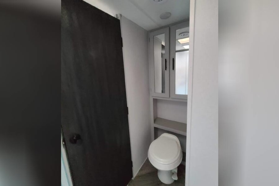 Inside an RV rental bathroom, featuring a white toilet, dark brown door, and mirrored cabinet above shelving. Functional space.