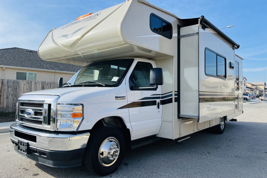A white and tan Coachmen Leprechaun Class C RV rental, on a Ford E-450 chassis, parked on a sunny street.