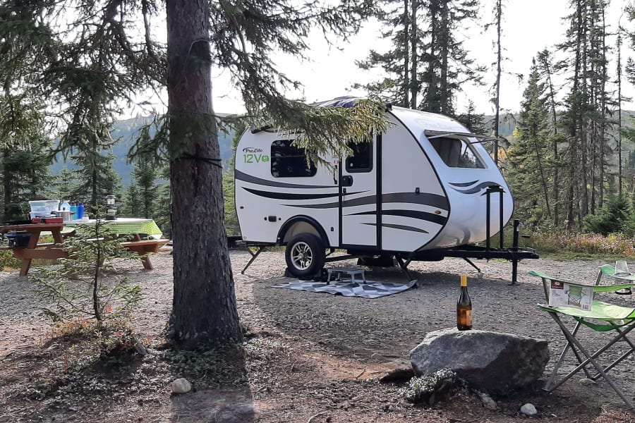 Peaceful nature escape! A compact white **rv rental** travel trailer is parked at a wooded campsite with a picnic table and green chairs.