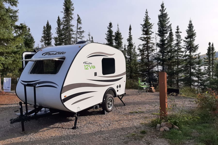 ProLite RV rental on a gravel campsite, surrounded by tall trees with a lake view. Ideal for outdoor travel and camping.