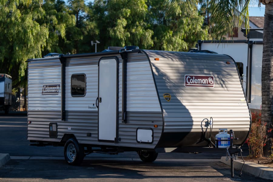 A compact gray and white Coleman travel trailer RV rental, parked on asphalt with a propane tank visible.