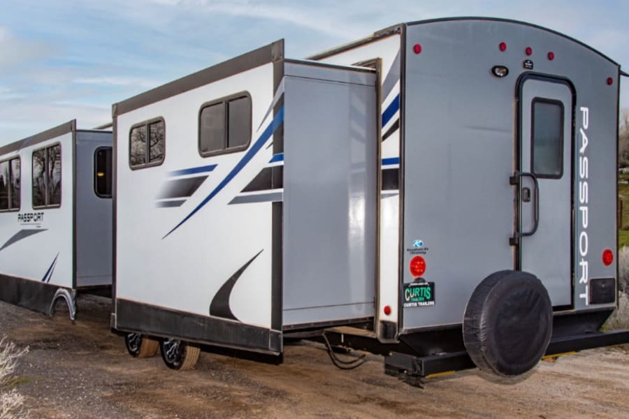A gray & white Passport travel trailer with extended slide-outs, parked outdoors, ideal for your rv rental.