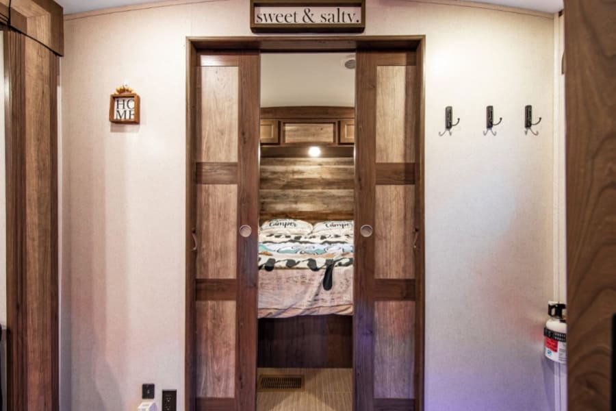 RV rental interior: Bedroom with wood sliding doors, a comfy bed, "sweet & salty" sign, and practical hooks.