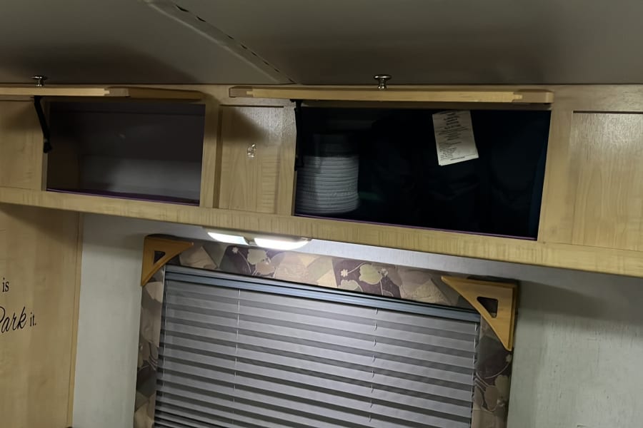 Cozy RV rental interior with wooden overhead storage cabinets, one open, a window with blinds, and a "Home is where you Park it" decal.