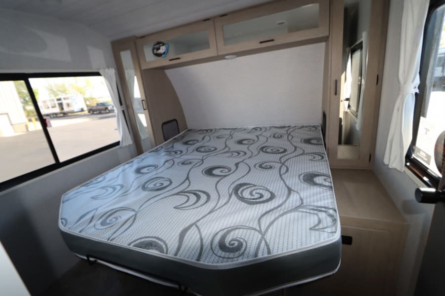 Compact RV rental bedroom featuring a bed with a patterned mattress, overhead cabinets, and large windows.