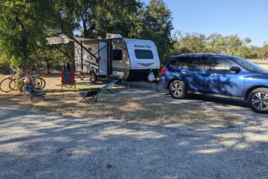 RV rental setup: A travel trailer with awning extended, SUV, bikes, and chairs at a sunny gravel campsite.