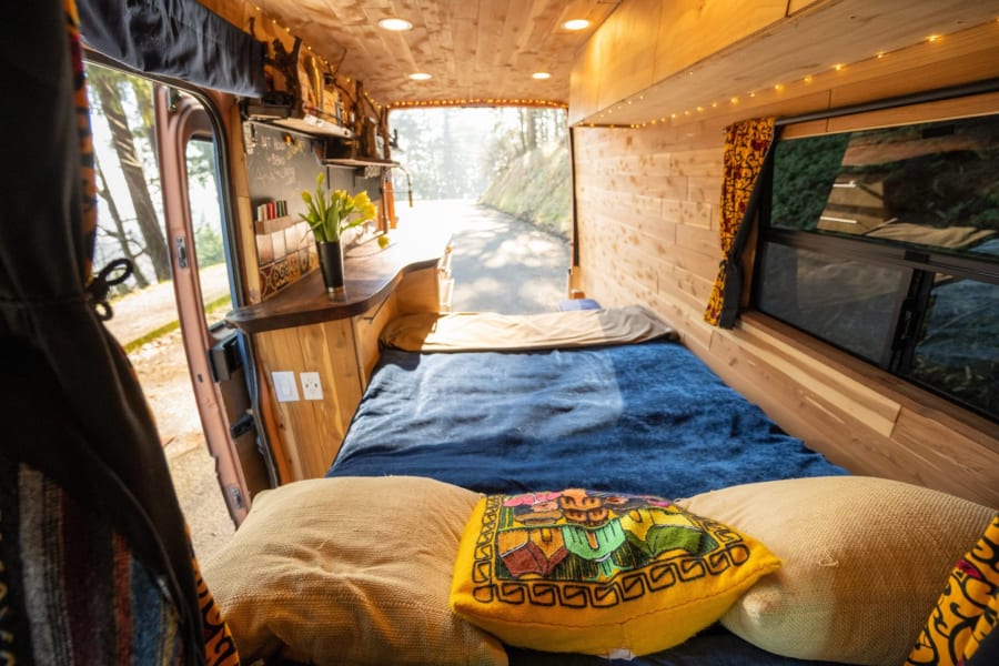 Experience van life! Cozy wood-paneled rv rental interior with bed & kitchen, opening to a scenic road for travel.