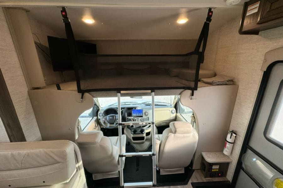 Inside an RV rental: View of the front overhead bunk with safety net, ladder, driver's cockpit, and nearby TV.