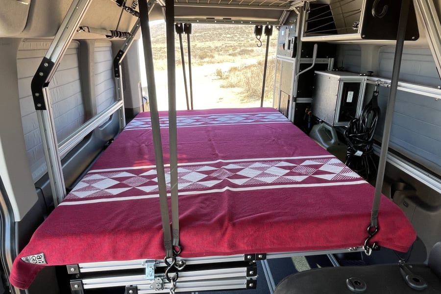 This RV rental showcases a cleverly suspended bed with a red blanket, modular storage, and scenic desert views.