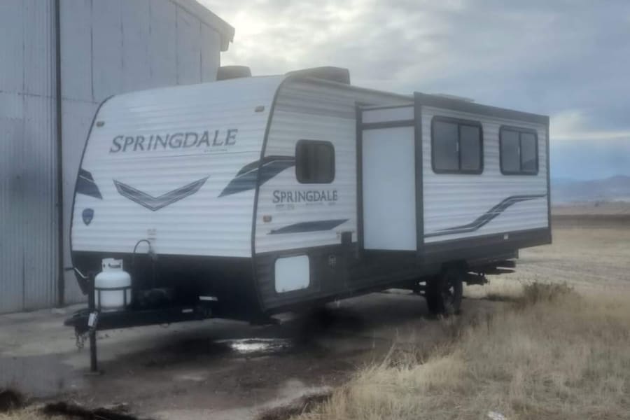 A Springdale RV rental travel trailer with slide-out extended, parked on muddy ground by a building under cloudy skies.