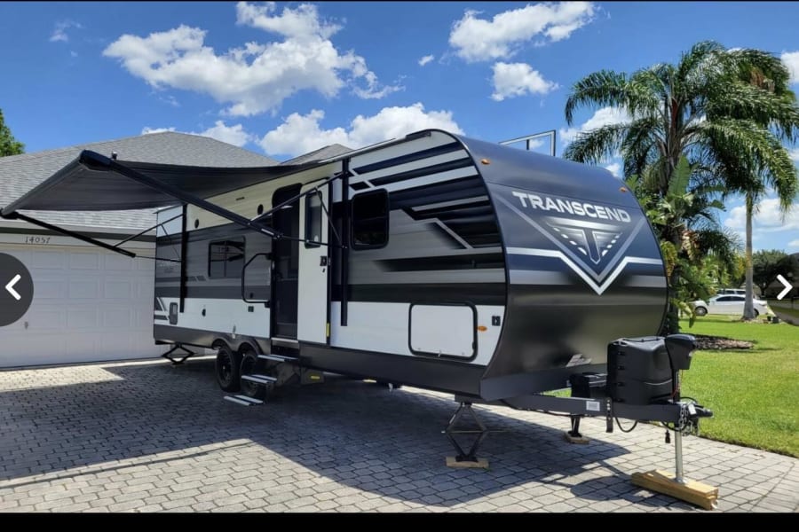 A sleek "TRANSCEND" travel trailer, ready for your next RV rental adventure, parked on a residential driveway with its awning extended.