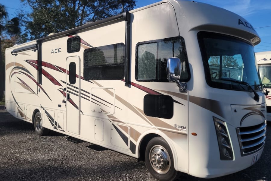 Spacious white Thor ACE Class A RV rental with brown/maroon graphics, seen from the front side on a gravel lot under sunny skies.