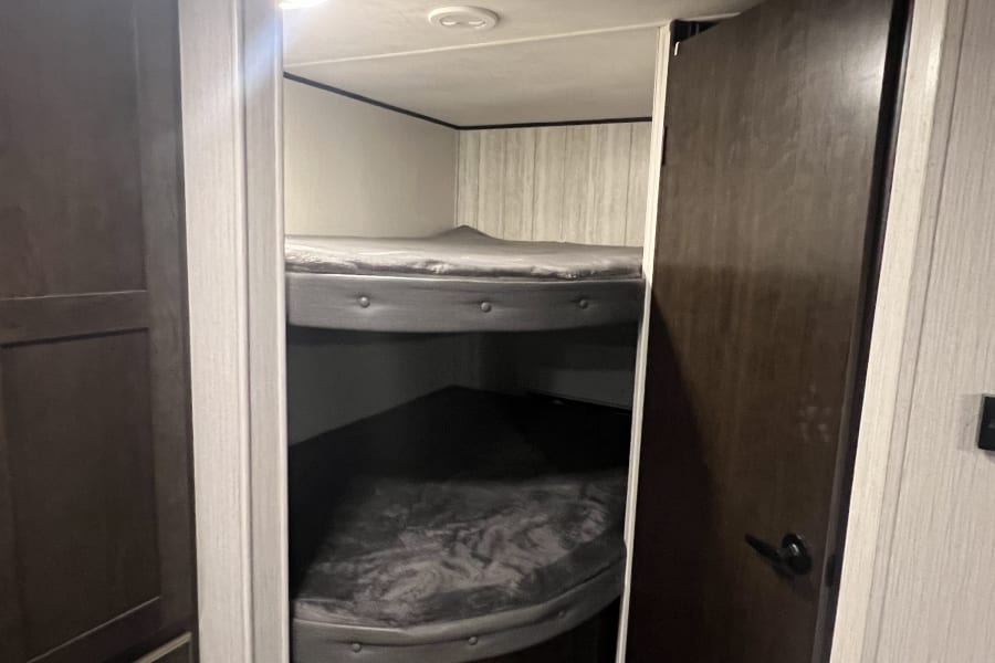 Compact RV rental interior: two cozy bunk beds with grey mattresses, dark wood storage, and bright lighting.