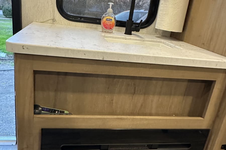 RV rental interior: a compact counter with sink, soap, paper towels, window view, control panel, and electric fireplace.