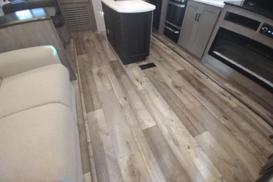 Explore this modern RV rental interior featuring a comfy sofa, wood-look floor, kitchen, and electric fireplace.