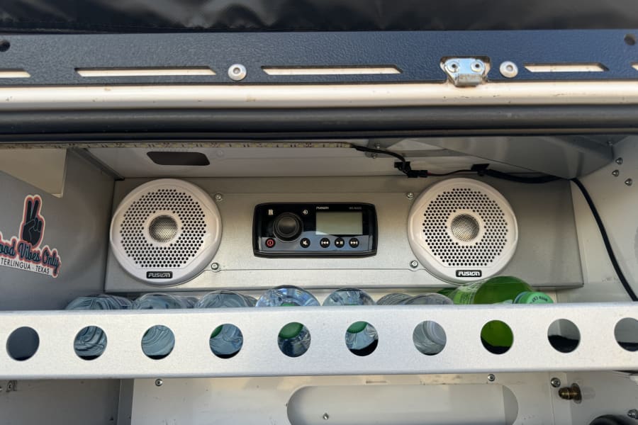 Custom RV rental setup features a Fusion stereo, speakers, and water bottles in a lit storage area. Good vibes!