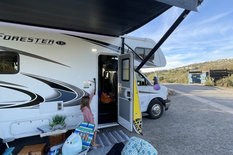 RV rental setup: A girl by a white RV with an extended awning, outdoor chairs, picnic basket, and a surfboard. Perfect for family camping.