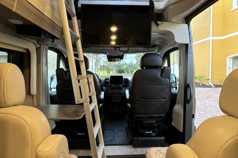Inside this rv rental: a full interior view from the rear, showing light tan captain's chairs, a bunk access ladder, and a mounted TV.