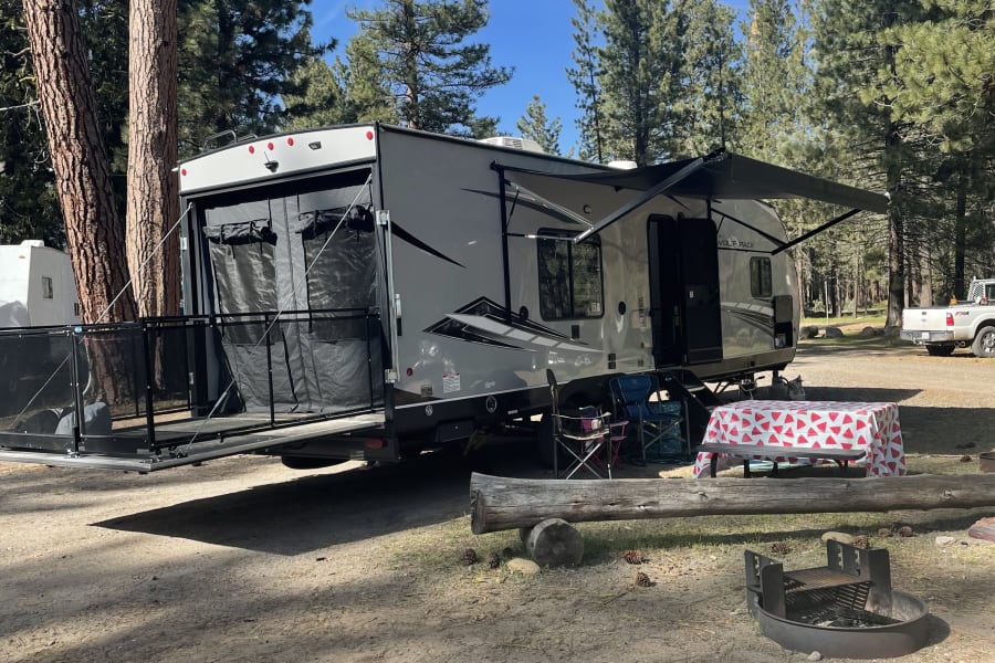 RV rental camping under tall pines. Features an awning, rear patio, fire pit, and picnic table with a watermelon tablecloth.