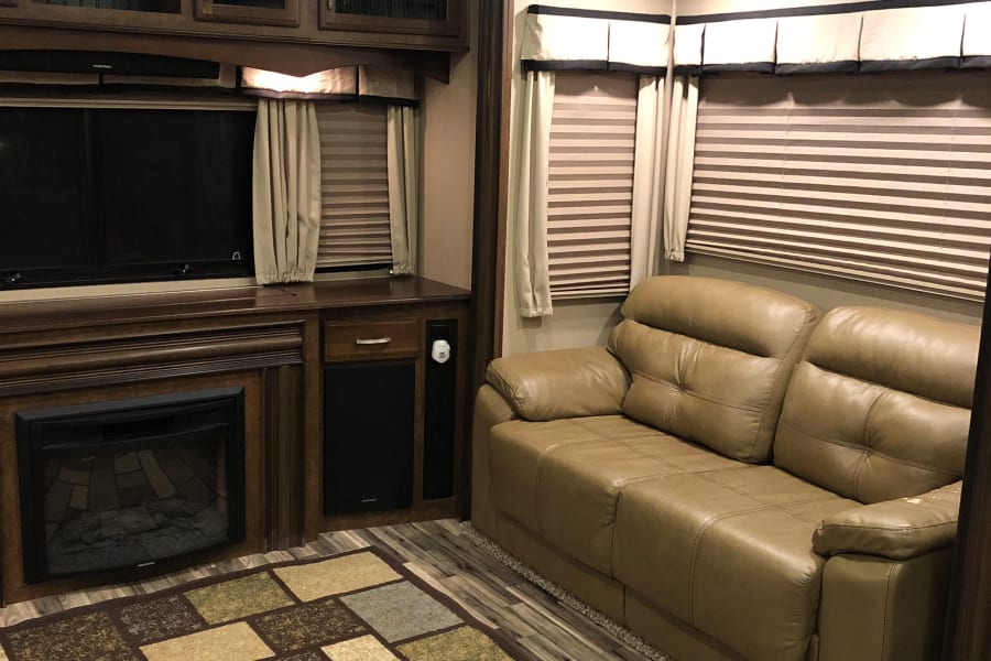 Step inside this inviting RV rental with a cozy brown leather sofa, electric fireplace, TV, and patterned rug in the living area.