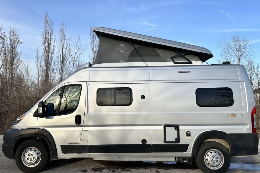 Silver Winnebago Solis camper van with pop-top for rv rental. Parked outdoors with bare trees & blue sky.