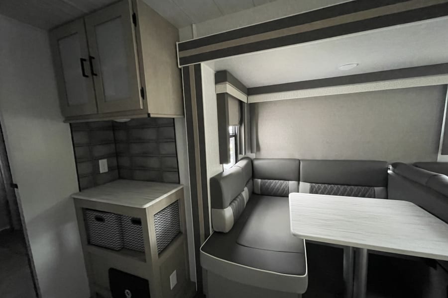 Inside a modern RV rental: a gray booth dinette with a white table, alongside a small kitchen counter with cabinets and storage.