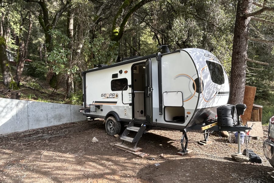 A GEO PRO RV rental is set up in a dirt camping area with a concrete wall, surrounded by lush trees.