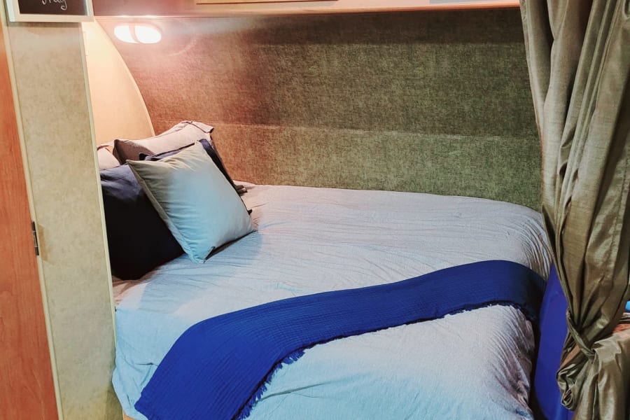 Cozy RV rental bedroom with blue bedding, overhead cabinets, a welcome sign, and a privacy curtain.