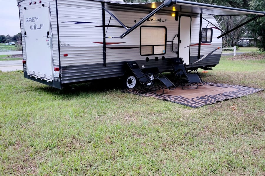 Grey Wolf travel trailer rv rental in a scenic outdoor setting, featuring an extended awning, mat, and three lounge chairs.