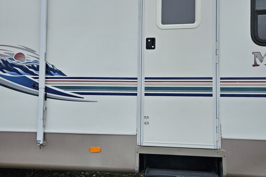 RV rental: Side of a white RV, highlighting the entry door, pull-out steps, and a retracted awning. Ground has grass/leaves.