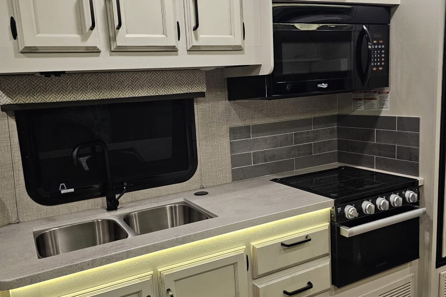 A bright, compact RV kitchen ready for your rv rental. Features white cabinets, stainless steel sink, microwave, and stove.