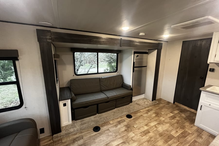 Inside a cozy RV rental: living area with sofa, large windows, refrigerator, and kitchenette. Perfect for travel.