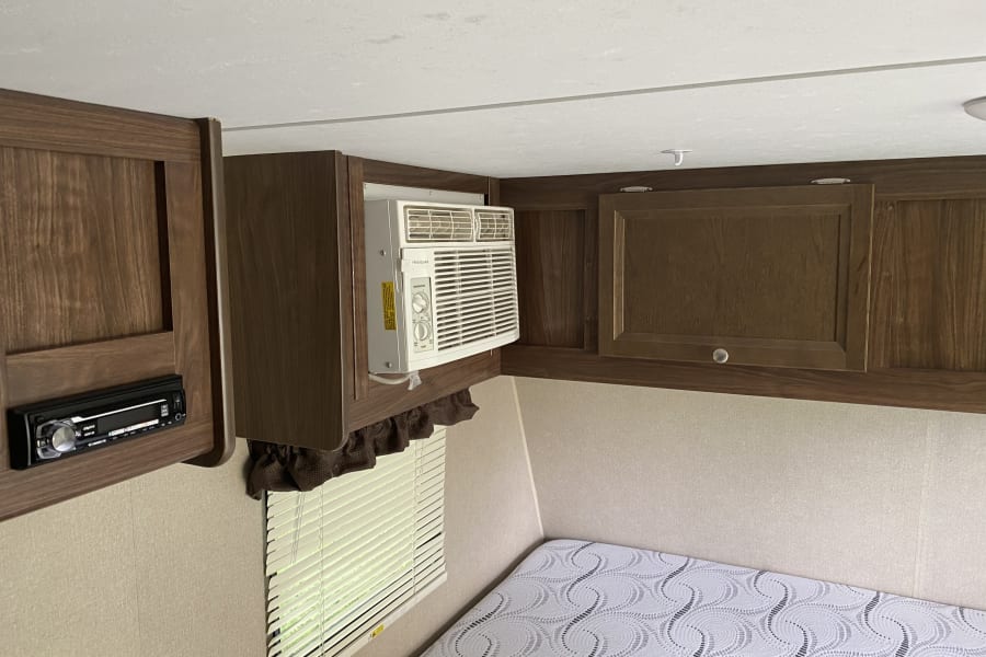 Inside an RV rental: a compact sleeping area with built-in wooden cabinets, a window AC, stereo, and a bed.