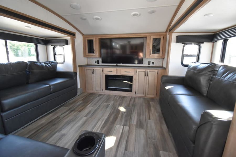 Spacious RV rental living room with two dark leather sofas facing a large TV and electric fireplace for comfortable travel.