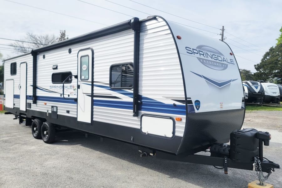 A white Springdale travel trailer with blue graphics, ideal for an RV rental or camping trip, parked on gravel.