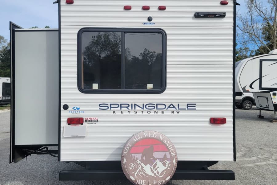 Rear view of a white Springdale Keystone travel trailer, ready for RV rental. Features a slide-out, window, and a "Not all who wander are lost" tire cover.