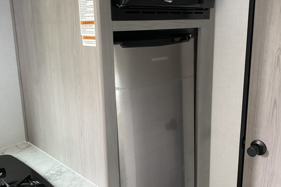 Compact RV rental kitchen features a black microwave, stainless Everchill fridge, and gas stovetop on a marble counter.