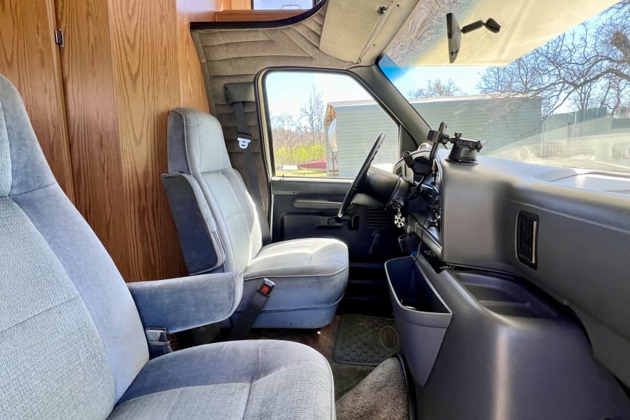 Explore this RV rental's cozy camper van interior. Features light blue fabric seats, wood cabinetry, and a spacious driving cabin.