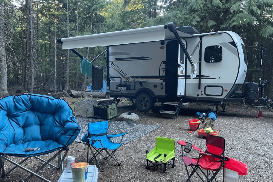 RV rental setup in a serene forest, featuring a white travel trailer, awning, and colorful camping chairs for an outdoor getaway.
