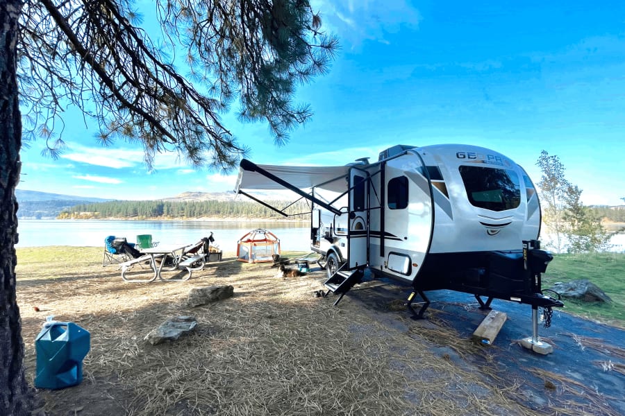Relaxing RV rental camping by the lake. A travel trailer, picnic table, chairs, and dogs are set up in this scenic spot.