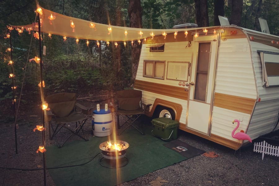 This charming rv rental scene features a camper, warm fire pit, flamingo lights, and chairs, nestled in nature for a cozy evening.