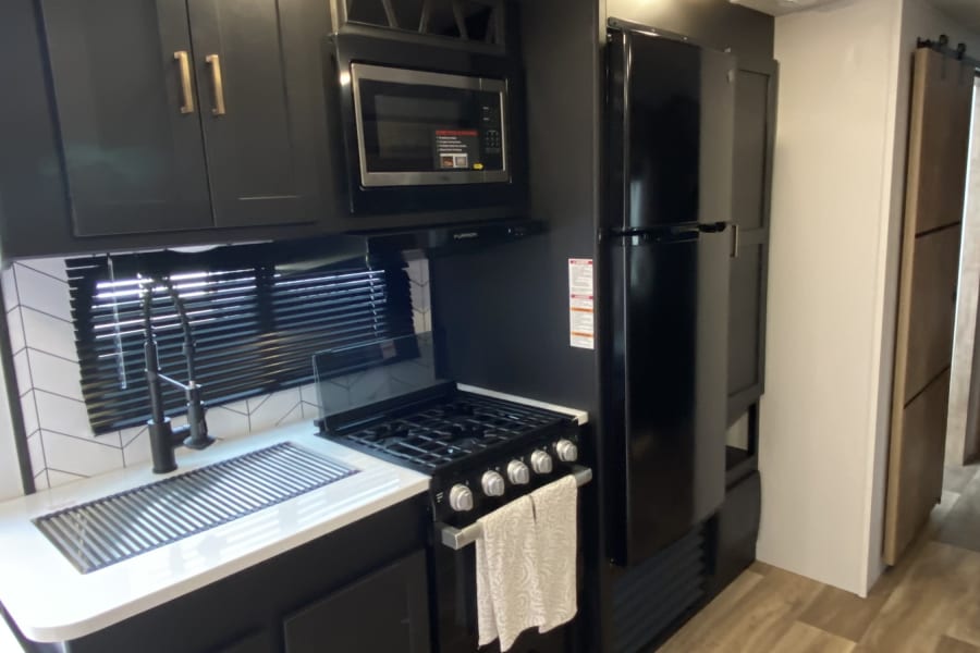 Inside an RV rental, this modern kitchen boasts black cabinets, a microwave, stove, sink, refrigerator, and light wood floors.